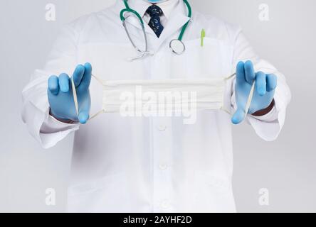 il medico in un cappotto bianco, i guanti sterili in lattice blu tiene in mano le maschere mediche in tessuto su uno sfondo bianco, maschera fatta in casa di garza bianca e garza Foto Stock