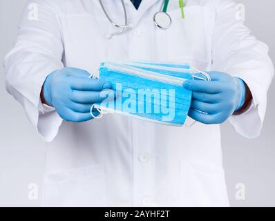 il medico in un cappotto bianco, i guanti sterili in lattice blu contengono le maschere mediche tessili in mano, gli accessori protettivi contro virus e batteri, da vicino Foto Stock