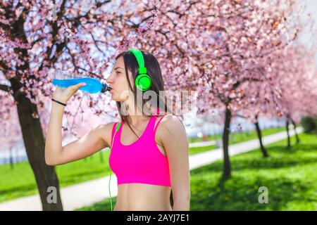 Bevanda energetica femminile durante l'allenamento. Ritratto di donna sportiva bevendo nel parco dopo il jogging. Adatta la musica d'ascolto della ragazza e gioita Foto Stock