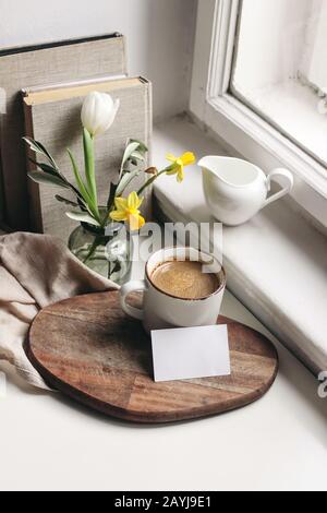 Accogliente primavera di Pasqua ancora vita. Scena di mockup di biglietto da visita. Tazza di caffè, libri, tagliere in legno, caraffa latte e vaso di narcisi e tulipano fl Foto Stock