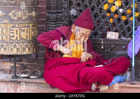 Vita in povertà povera Vecchia donna nepalese in rosso vestito e rugosa faccia seduta e implorante da ruota di preghiera buddista a Kathmandu, Nepal Foto Stock