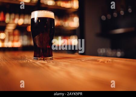 Bicchiere di birra scura artigianale con schiuma su bancone bar in legno, pub sfondo sfocato Foto Stock