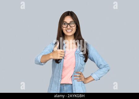 Felice giovane ragazza indiana guardare la fotocamera mostra pollici su sfondo grigio studio Foto Stock