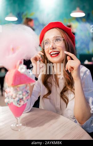 Francia dolce dessert nazionale, ragazza in beretto rosso mangia torta rosa, caramella floss e milkshake, ride e sorrisi Foto Stock