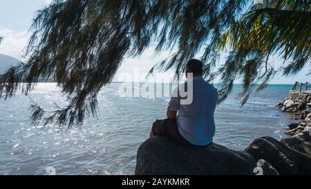 Lungo l'uomo seduto su una roccia rilassante e riflessione con un bel cielo blu e mare Foto Stock