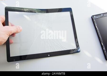 Un tecnico tiene in mano il vetro rotto dello schermo a sfioramento di un computer tablet rimosso per la sostituzione. Riparazione di apparecchiature elettroniche e configurazioni Foto Stock