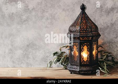 Marocchino ornamentale nero, lanterna araba. Foglie di olive verdi, rami su vecchio tavolo di legno, sfondo muggato muro,. Biglietto d'auguri per musulmani Foto Stock