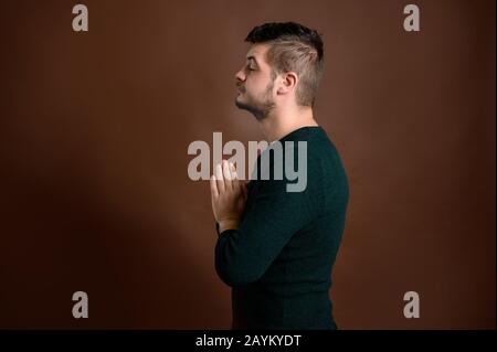 Giovane uomo con capelli castani vestito casualmente vista laterale è pregando mano aggrappato sperando per il perdono su sfondo marrone isolato Foto Stock