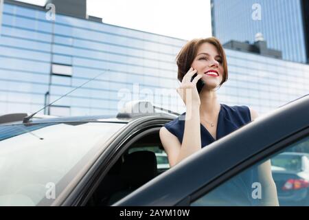 donna di affari con le labbra rosse in un vestito esce dalla macchina e parla al telefono. Foto Stock