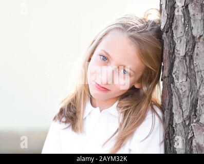 Una bella ragazza bionda dai capelli biondi, con occhi blu e un sorriso penoso sul suo viso, indossando una camicia bianca, appoggiata sul tronco di una pineta Foto Stock