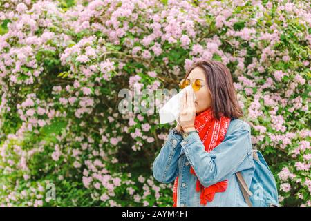 Concetto di allergia. Giovane donna asiatica starnutisce e soffia il suo naso con un fazzoletto e la sofferenza in primavera tra alberi fioriti e fioriti. Foto Stock