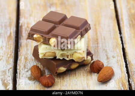 Pezzi di cioccolato bianco e latte con mandorle e nocciole impilati su superficie di legno, primo piano Foto Stock