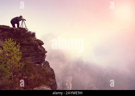 Fotografo sulla scogliera di montagna che guarda la nebbia attraverso il mirino. L'escursionista farà scattare foto con la fotocamera in mano. Foto Stock