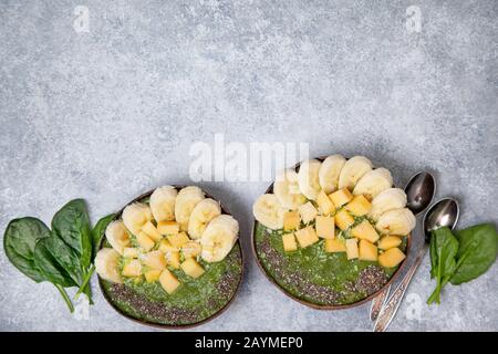 ciotola di spinaci frullato con banana e mango, semi di chia in ciotole di cocco su fondo grigio di cemento. vista dall'alto. Foto Stock