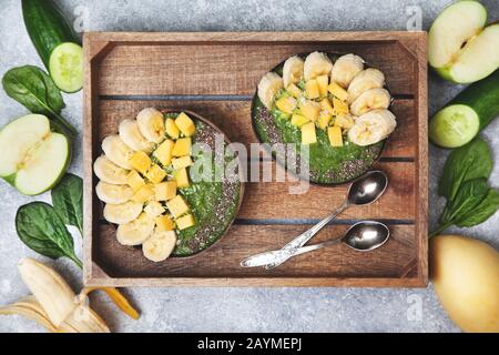ciotola di spinaci frullato con banana e mango, semi di chia in ciotole di cocco su fondo grigio di cemento. vista dall'alto. Foto Stock