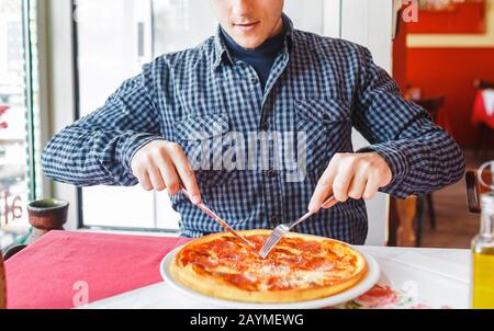 Un uomo affamato che mangia la pizza con forchetta e coltello nel ristorante italiano Foto Stock