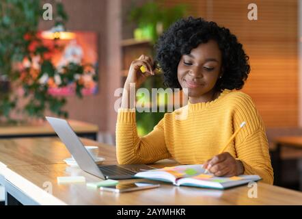 Ragazza nera sognante che scrive giù i pensieri, spendendo il tempo al caffè Foto Stock