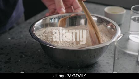 uomo che mescola ingredienti secchi con farina in ciotola d'acciaio su ripiano in cemento, foto larga Foto Stock