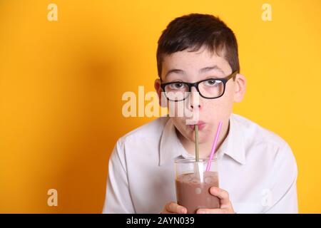 Un ragazzo giovane che indossa bicchieri di 8yrs vecchio beve un bicchiere di latte al cioccolato su sfondo giallo Foto Stock