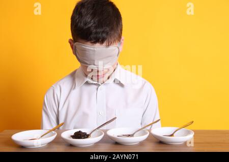 Prova del gusto alla cieca, ragazzo di 12 anni seduto con piatti di cibo campione per degustazione diversi prodotti alimentari bendati Foto Stock