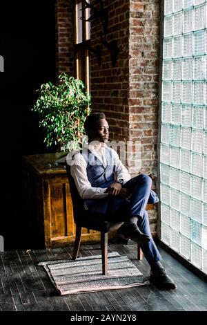 Giovane uomo d'affari con pelle scura che pensa al suo lavoro comodamente seduto davanti alla finestra Foto Stock