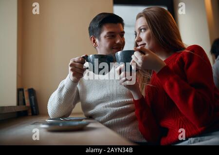 La coppia dell'ippopotamo il caffè yummy, seduta vicino a vicenda, guardandosi l'altro l'uomo sta portando un mentre maglione e la donna sta portando re Foto Stock