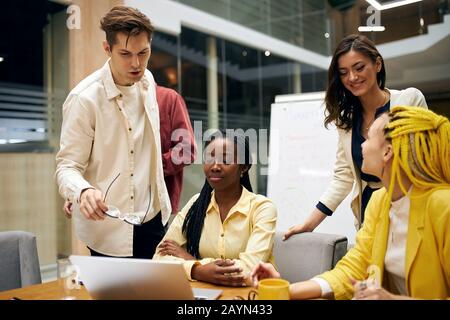 Giovane uomo in maglietta bianca che dà una mano d'aiuto al nuovo lavoratore coafricano che è seduto al tavolo di fronte al laptop.help , consiglio Foto Stock