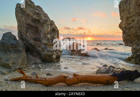 Tramonto tra grandi rocce, onde lattiche e grande tronco in primo piano, lunga esposizione, colori caldi, famosa Kathisma Beach, Lefkada, Grecia Foto Stock