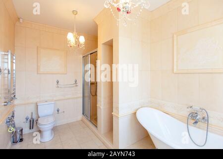 Bagno classico in toni beige. Vasca da bagno curva. Grande piastrella beige. Piastrelle bianche sul pavimento. Lampadario ritorto, elegante e costoso impianto idraulico Foto Stock