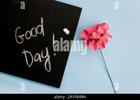 Concepimento Buongiorno. Bordo di gesso nero con l'iscrizione buon giorno, fiore rosa su uno sfondo blu chiaro. Vista dall'alto. Disposizione piatta Foto Stock