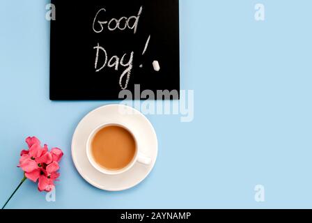 Concepimento Buongiorno. Lavagna in gesso nero con l'iscrizione buon giorno, una tazza di caffè rosa su uno sfondo blu chiaro. Vista dall'alto. Disposizione piatta. Foto Stock