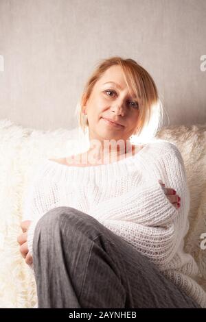 Una bella donna adulta è seduta in una stanza su un divano. Ritratto di una bionda a casa. Foto Stock