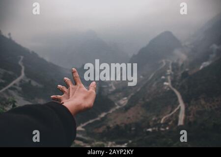 Una mano che raggiunge le nebbie montagne e le strade tortuose di ha giang Loop in Vietnam mostrando una fotografia di viaggio cinematografica e moody Foto Stock