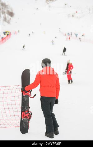 uomo in casco nero che indossa lo sport invernale che tiene snowboard guardando la montagna di neve con sciatori e snowboarder giovane ragazza in vestiti rossi Foto Stock