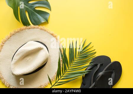 Cappello da spiaggia, infradito e foglie di monstera. Sfondo estivo con accessori gialli. Vista dall'alto, piatto. Spazio per il testo. Foto Stock