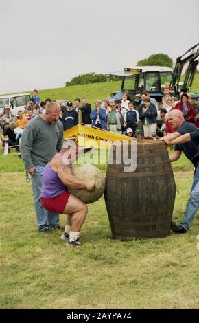 1980s, 'gara uomo più forte'...fuori in un campo d'erba guardato dagli spettatori, un concorrente maschile che solleva una palla di pietra pesante o una sfera da terra con l'obiettivo di sollevarla sulla cima di un vecchio barile di whisky di legno in un concorso uomo più forte, Highlands, Scozia, Regno Unito. L'evento può essere valutato sia sul peso della pietra che sul tempo preso o su entrambi. Foto Stock