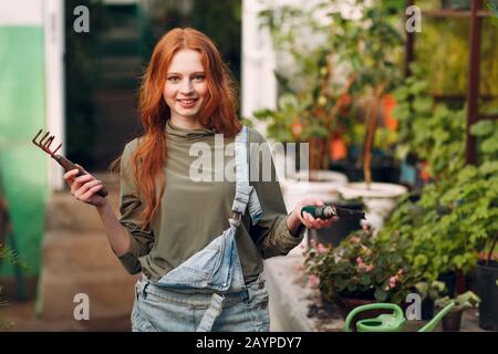 Concetto di giardinaggio domestico. Giovane agronomo zenzero donna in tute denim e rastrello e pala piantare piante. Primavera casa giardino pianta. Foto Stock