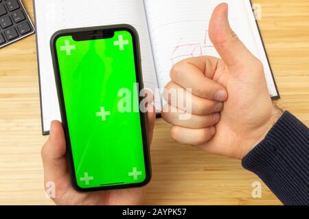 Immagine mockup del telefono cellulare nero con schermo verde sul tavolo dell'ufficio sullo sfondo con strumenti di cancelleria. Una mano d'uomo mostra come Foto Stock