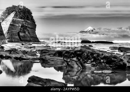 Bellissime formazioni rocciose sulla spiaggia con il cono vulcanico del Monte Egmont in lontananza Foto Stock