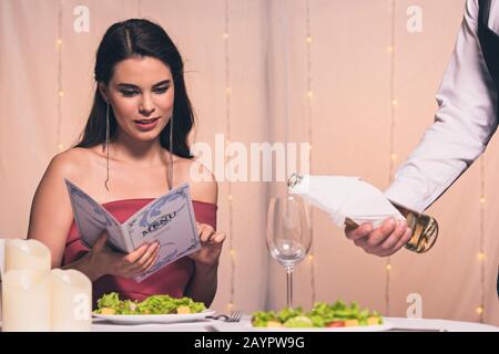 attraente ed elegante menu da lettura per le ragazze mentre il cameriere versa vino bianco in vetro Foto Stock