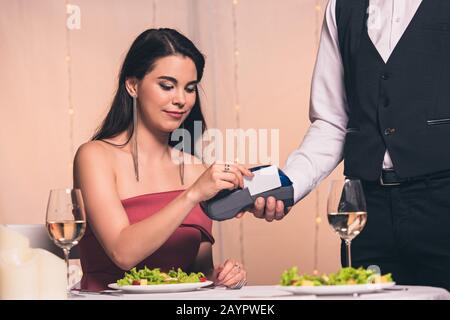 vista ritagliata del cameriere che tiene il terminale di pagamento vicino bella, ragazza elegante seduto al tavolo servito Foto Stock