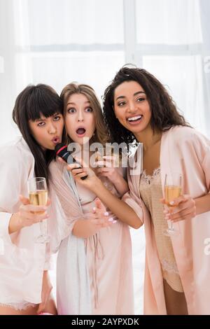 belle ragazze multiculturali eccitate che tengono champagne e cantano con il microfono sulla festa del bachelorette Foto Stock