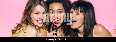 scatto panoramico di belle ragazze multietniche felici che cantano karaoke con microfono, isolato su rosa Foto Stock