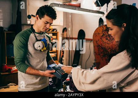 lavoratore che tiene il terminale e donna sorridente che paga con carta di credito e che tiene snowboard nel negozio di riparazione Foto Stock