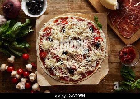 Vista dall'alto della pizza su carta pergamena con parmigiano, verdure, olive e salame su fondo di legno Foto Stock
