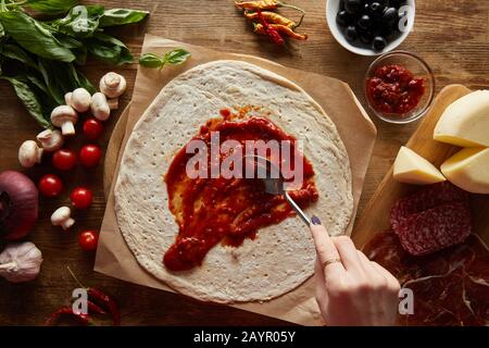 vista corta della donna che aggiunge salsa di pomodoro sulla pasta vicino agli ingredienti per la pizza su tavola di legno Foto Stock