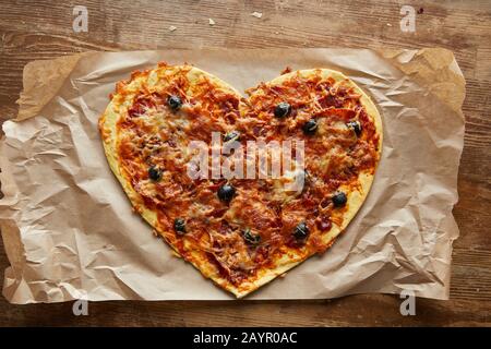 vista dall'alto della deliziosa pizza italiana a forma di cuore sulla carta da forno su un tavolo in legno Foto Stock