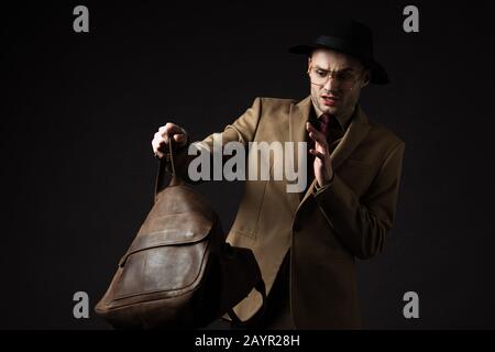 uomo elegante e stressato in abito beige, cappello e occhiali che guarda in borsa di pelle marrone isolato su nero Foto Stock