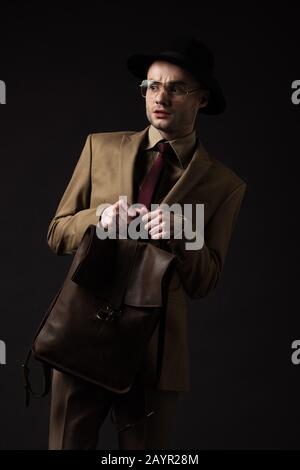 uomo elegante preoccupato in abito beige, cappello e occhiali che tengono zaino in pelle marrone isolato su nero Foto Stock