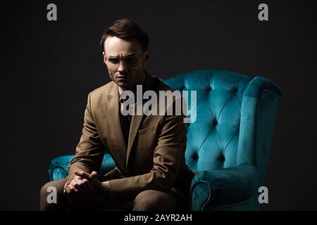 uomo elegante teso in abito beige occhiali seduti con mani stringate in velluto blu poltrona isolato su nero Foto Stock